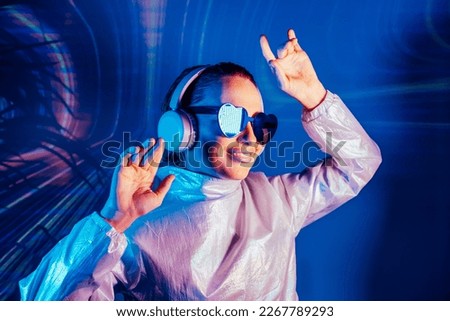 Portrait of a smiling dancing woman in sunglasses and headphones in neon light. Music lover. Silent disco. Woman in holographic clothes on the background of music vibes background. Futuristic Party.