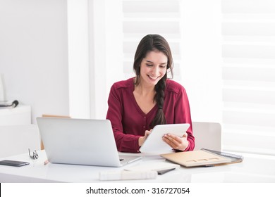 Portrait Of A Smiling Dark Braided Hair Woman Home Working In Casual Clothes. She Is Sitting At A Big White Table Holding Her Tablet And Looking At Her Laptop And Some Documents In Front Of Her