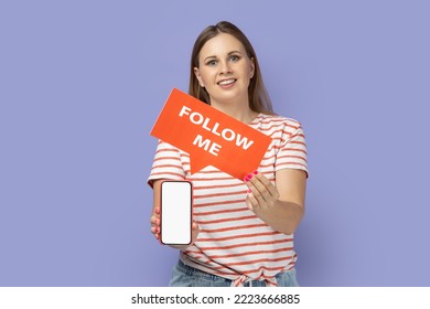Portrait of smiling delighted blond woman wearing striped T-shirt holding red card with follow me inscription and showing white empty display of phone. Indoor studio shot isolated on purple background