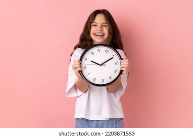 Portrait of smiling delighted little girl wearing white T-shirt holding wall clock, being happy, deadline, satisfied with completed home task. Indoor studio shot isolated on pink background. - Powered by Shutterstock