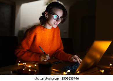 Portrait Of Smiling Designer Girl, Working At Graphic Tablet On Laptop. Wearing Eyeglasses And Orange Sweater. In Dark Room Home, With Garlands.