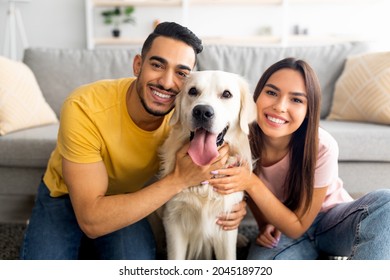 Portrait of smiling diverse couple hugging their pet dog, smiling at camera, indoors. Arab guy and his Caucasian girlfriend embracing their golden retriever at home - Powered by Shutterstock