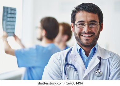 Portrait Of Smiling Doctor Looking At Camera On Background Of His Working Colleagues