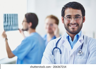 Portrait Of Smiling Doctor Looking At Camera On Background Of His Working Colleagues