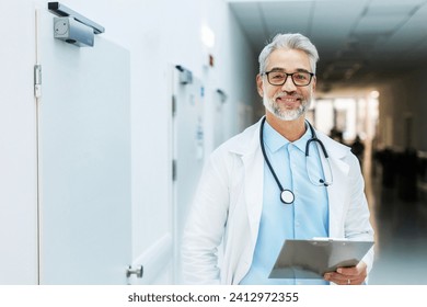 Portrait of smiling doctor standing in hospital corridor. Handsome doctor with gray hair wearing white coat, stethoscope around neck standing in modern private clinic, looking at camera. - Powered by Shutterstock