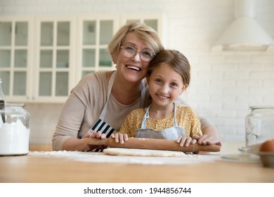 Portrait of smiling elderly 60s Caucasian grandmother and little 8s granddaughter baking together in home kitchen. Happy mature 50s granny and small grandchild cooking pie or cookies on weekend.