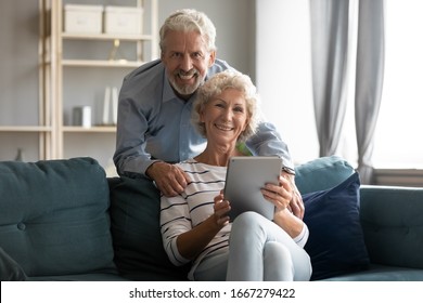 Portrait Of Smiling Elderly 60s Couple Sit Relax On Cozy Couch In Living Room Using Modern Tablet Together, Happy Old Middle-aged 50s Husband And Wife Look At Camera Browsing Surfing Internet On Pad