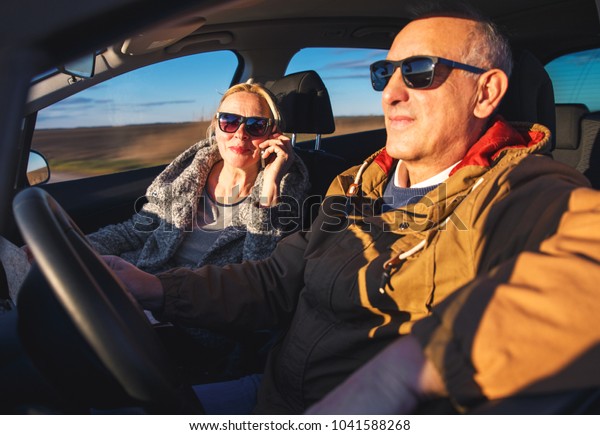 Portrait of smiling elderly couple\
driving car, while female passenger talking on\
phone.