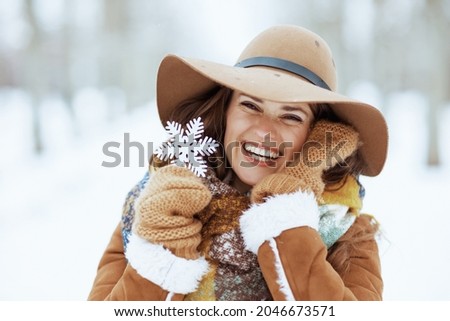 Portrait of smiling elegant 40 years old woman in brown hat and scarf with mittens and snowflake in sheepskin coat outside in the city park in winter.