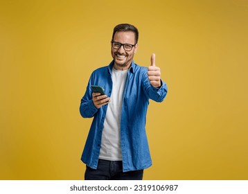 Portrait of smiling entrepreneur showing thumbs up and messaging on smartphone on yellow background