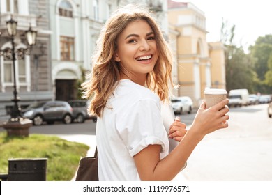 Portrait of smiling european woman strolling through city street with silver laptop and takeaway coffee in hands - Powered by Shutterstock