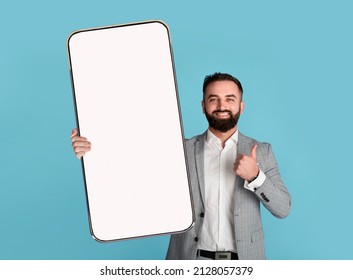 Portrait Of Smiling Excited Bearded Business Man In Suit Holding Big Smartphone With White Empty Screen In Hand, Showing Thumbs Up Gesture. Free Copy Space For Mock Up, Banner, Blue Studio Background