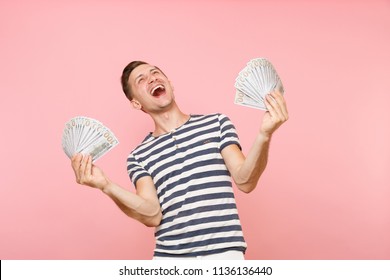 Portrait Of Smiling Excited Young Man In Striped T-shirt Holding Bundle Lots Of Dollars, Cash Money, Ardor Gesture On Copy Space Isolated On Pink Background. People Sincere Emotions Lifestyle Concept