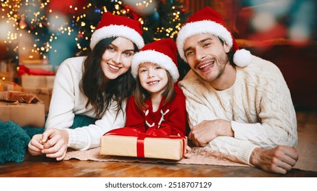 Portrait of smiling family in Santa hats laying on floor by xmas tree, daughter holding gift - Powered by Shutterstock