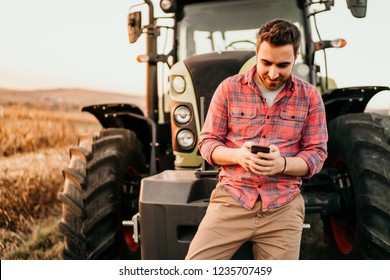 Portrait Of Smiling Farmer Using Smartphone And Tractor At Harvesting. Modern Agriculture With Technology And Machinery Concept