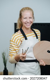 Portrait Of A Smiling Female Art Teacher. She's Wearing Apron, Holding Wooden Palette And A Bundle Of Brushes.