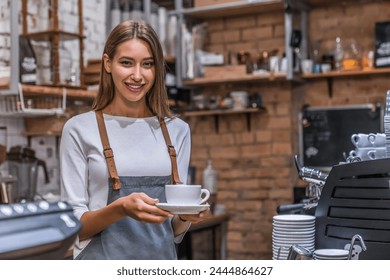 Portrait of smiling female barista holding cup with coffee at coffee shop. Beautiful young woman waitress serving coffee tea cappuccino with a big smile. Service industry. Small business concept - Powered by Shutterstock