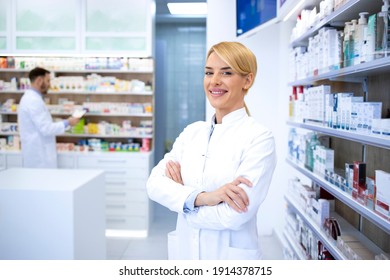 Portrait of smiling female blonde pharmacist standing in pharmacy shop or drugstore with her arms crossed. In background shelves with medicines.