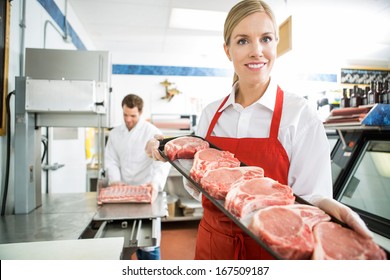 Portrait of smiling female butcher holding meat tray at store with colleague working in background - Powered by Shutterstock