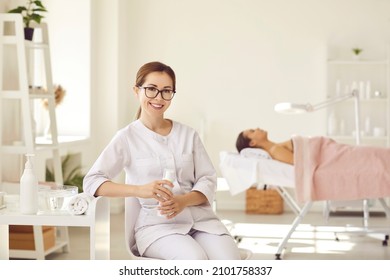 Portrait Of Smiling Female Dermatologist, Skin Therapist, Beautician And Aesthetic Professional. Happy Beautiful Woman In Clean White Coat Uniform And Glasses Sitting In Modern Interior Of Her Office