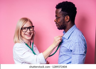 Portrait of a smiling female doctor with afro american patient man.beautiful blonde woman white medical coat and glasses holding a stethoscope on the heart the sick on a pink background in the studio.
