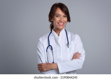 Portrait Of Smiling Female Doctor With Arms Crossed Standing Against At Isolated Grey Background. Confident Healthcare Worker Is Wearing Lab Coat. 