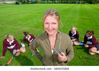 Portrait of smiling female field hockey coach holding the whistle in her hand with the blurred background of girls sitting on the ground. - Powered by Shutterstock