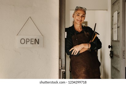 Portrait of smiling female jeweler in apron with tools standing at workshop door. Senior jeweler in apron holding tools at workshop. - Powered by Shutterstock