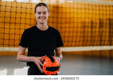 Portrait of a smiling female professional volleyball player posing while holding a ball while playing a match or training in a sports hall. Woman standing in front of a volleyball net. Copy space. - Powered by Shutterstock