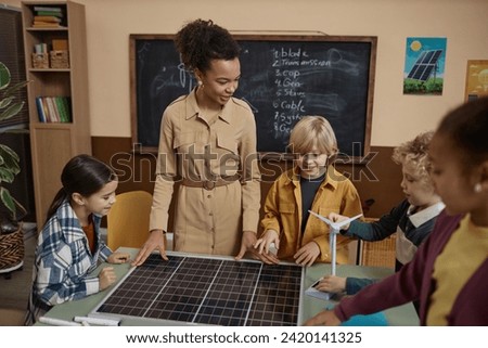 Portrait of smiling female teacher showing solar panel to class of little children learning about eco friendly energy in school