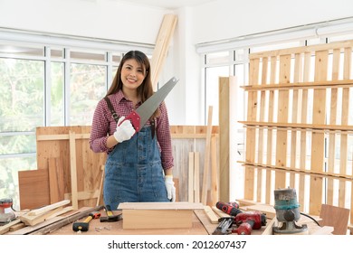 Portrait Of Smiling Females Carpenter Holding A Hand Saw On Wooden Background In Carpentry Shop. Asian Handywoman Apprentice Working In The Workshop. DIY Woodworking Crafts And Hobbies Concepts. 