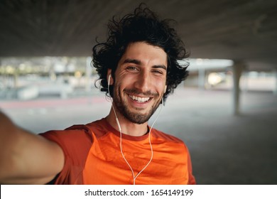 Portrait of a smiling fit young man with earphones in his ears taking selfie outdoors - pov shot of a man looking at the camera smiling taking a selfie