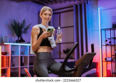 Portrait of smiling fitness girl using mobile phone and drink water after training on exercise bike in evening at modern home. Portrait of model, fit sporty woman, at home engaged in a stationary bike - Powered by Shutterstock