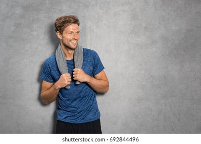 Portrait of a smiling fitness man with towel on shoulders looking away. Happy young man relaxing after training and leaning on grey wall. Sporty active guy resting after workout with copy space.