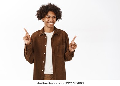 Portrait Of Smiling Gen Z Guy With Afro Hair, Pointing Fingers Sideways, Showing Left And Right Side, Logo Or Product Banners, Give Choice, Standing Over White Background