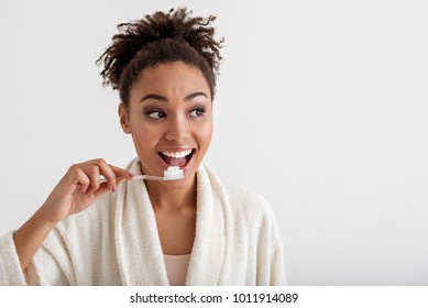 Portrait of smiling girl brushing teeth. She is wearing housecoat and looking aside with happiness. Copy space in right side. Isolated on background
