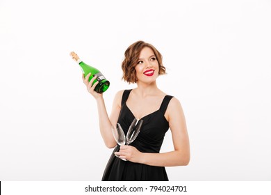 Portrait of a smiling girl dressed in black dress holding bottle of champagne and two glasses isolated over white background