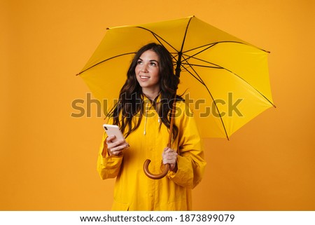 Portrait of a smiling girl dressed in raincoat standing with an open umbrella and using mobile phone isolated over yellow background