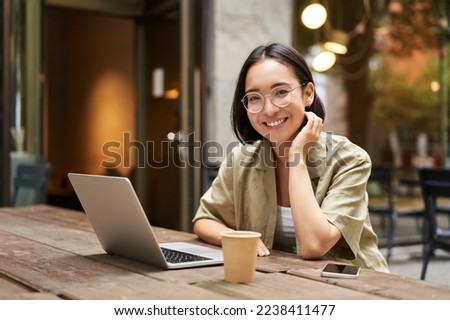 Portrait of smiling girl in glasses, sitting with laptop in outdoor cafe, drinking coffee and working remotely, studying online.
