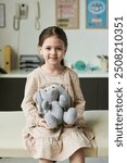 Portrait of smiling girl holding a gray stuffed animal in a hospital room, sitting on examination table and looking at camera. Background includes medical equipment and posters