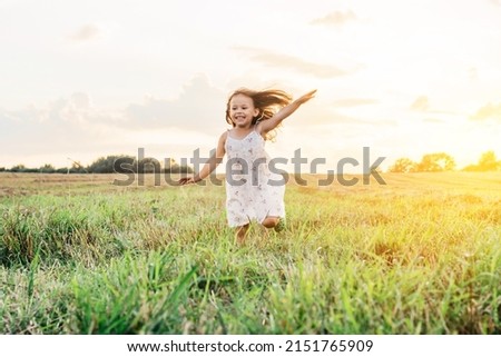 Portrait of smiling girl playing, jumping and running on grass hay field paths of dry grass in the sunset. Waving hands. Forest on bright light background. Cloudy sunny sky. Haying time
