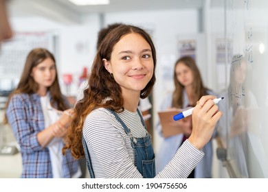 Portrait of smiling girl solving math equation on white board. College student thinking and solving arithmetic sum with classmates. Smart young woman writing on white board during class.