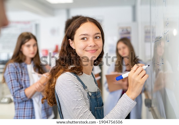 Portrait\
of smiling girl solving math equation on white board. College\
student thinking and solving arithmetic sum with classmates. Smart\
young woman writing on white board during\
class.