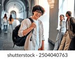 Portrait of a smiling handsome curly-haired student in glasses with a backpack standing in the corridor of a university building. Ready to study, student life.   