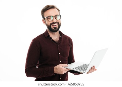 Portrait of a smiling handsome man in eyeglasses holding laptop computer and looking at camera isolated over white background