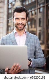 Portrait of a smiling handsome man in jacket using laptop computer and looking at camera while standing in a city area - Shutterstock ID 658230181