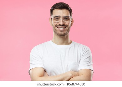 Portrait of smiling handsome man in white t-shirt and transparent eyeglasses standing with crossed arms isolated on pink background