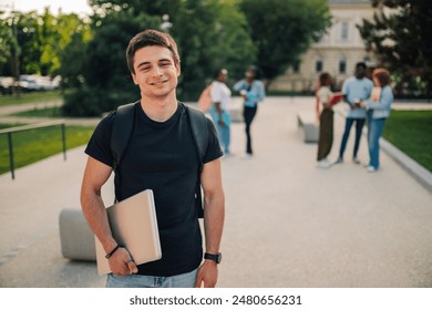 Portrait of smiling handsome university student boy standing at college campus with laptop in hand and looking at camera. Academic lifestyle. Adolescent college student posing at university campus. - Powered by Shutterstock