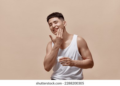 Portrait of smiling, handsome, young guy with clean, spotless face applying face moisturizing cream against light brown studio background. Concept of male beauty, skincare, cosmetology, men's health - Powered by Shutterstock