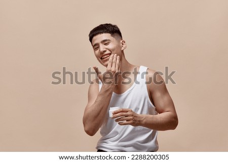 Portrait of smiling, handsome, young guy with clean, spotless face applying face moisturizing cream against light brown studio background. Concept of male beauty, skincare, cosmetology, men's health